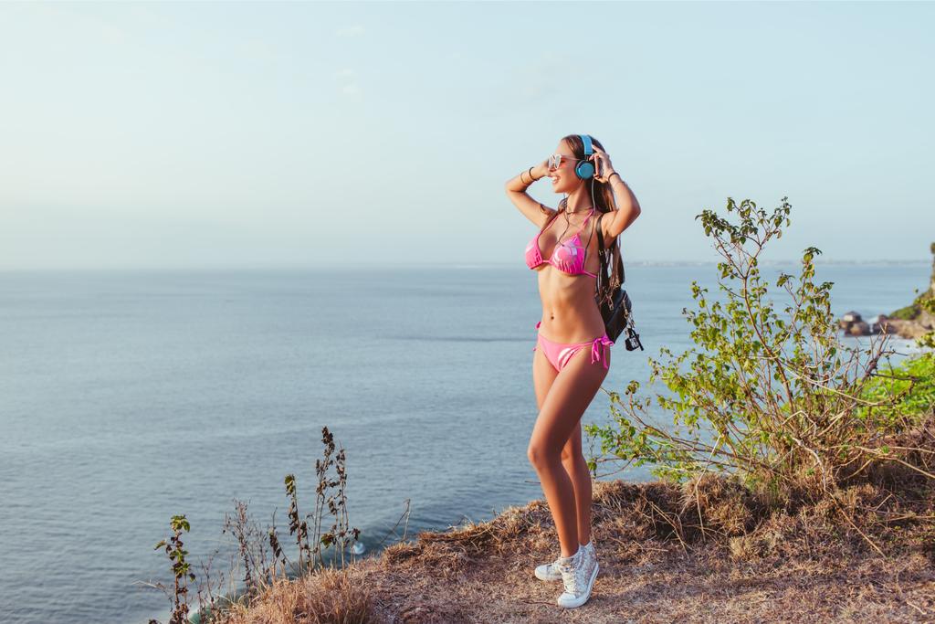 chica atractiva en bikini rosa tocando los auriculares y mirando a la playa
 - Foto, imagen