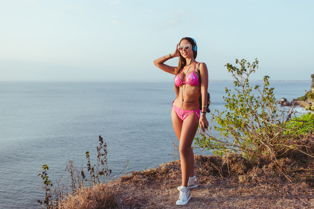 Улыбающаяся привлекательная девушка в наушниках и розовом бикини, стоящая на берегу моря
 - Фото, изображение