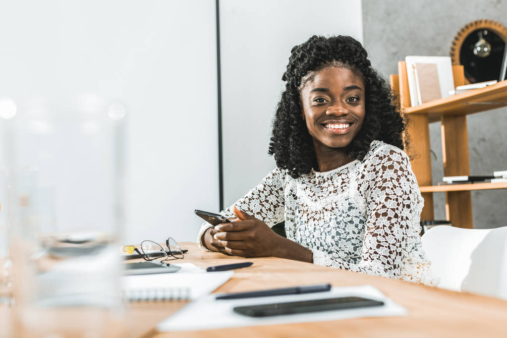 портрет усміхненої афроамериканської бізнес-леді зі смартфоном в руках дивиться на камеру, сидячи на робочому місці
 - Фото, зображення