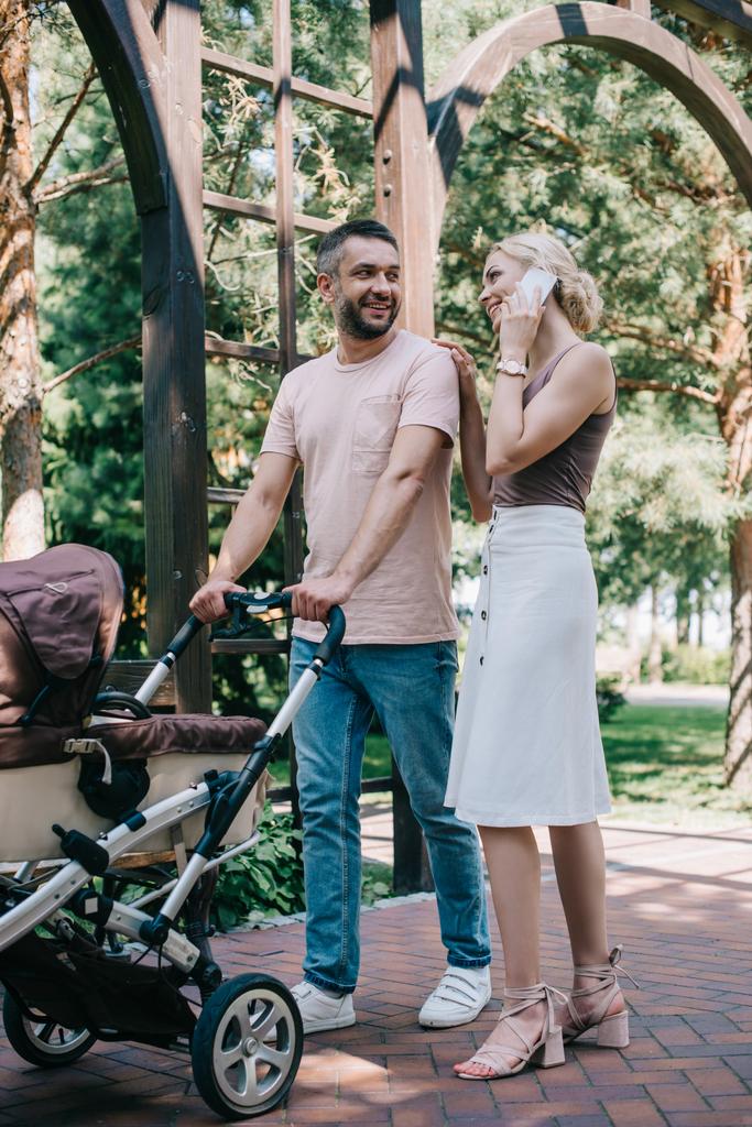 šťastný matka mluví o smartphone poblíž kočárek a palming manžel v parku - Fotografie, Obrázek