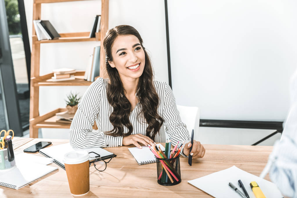 портрет улыбающейся азиатской предпринимательницы, сидящей на рабочем месте с различными офисными принадлежностями
 - Фото, изображение