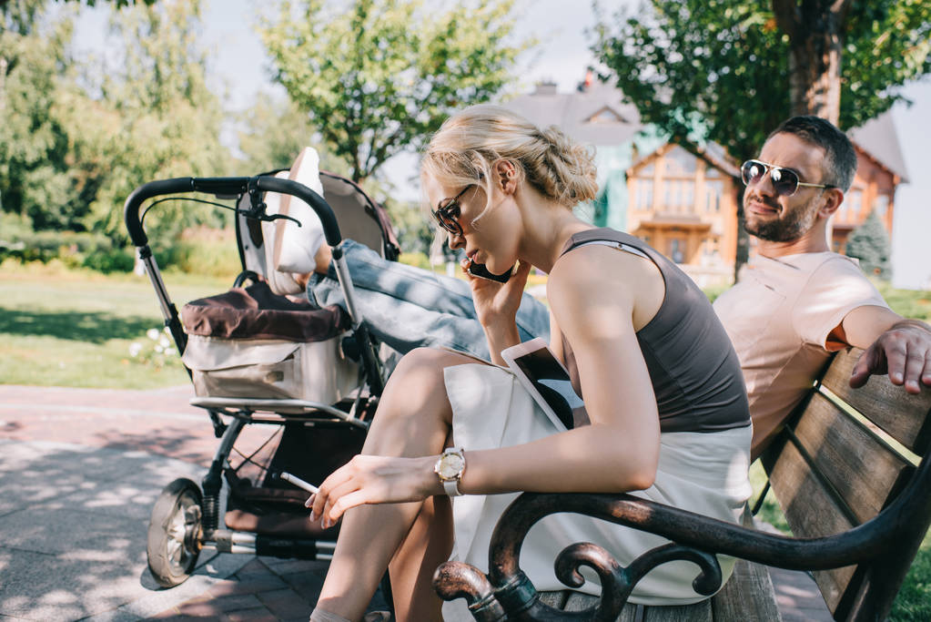 mother smoking cigarette near baby carriage in park and talking by smartphone - Photo, Image
