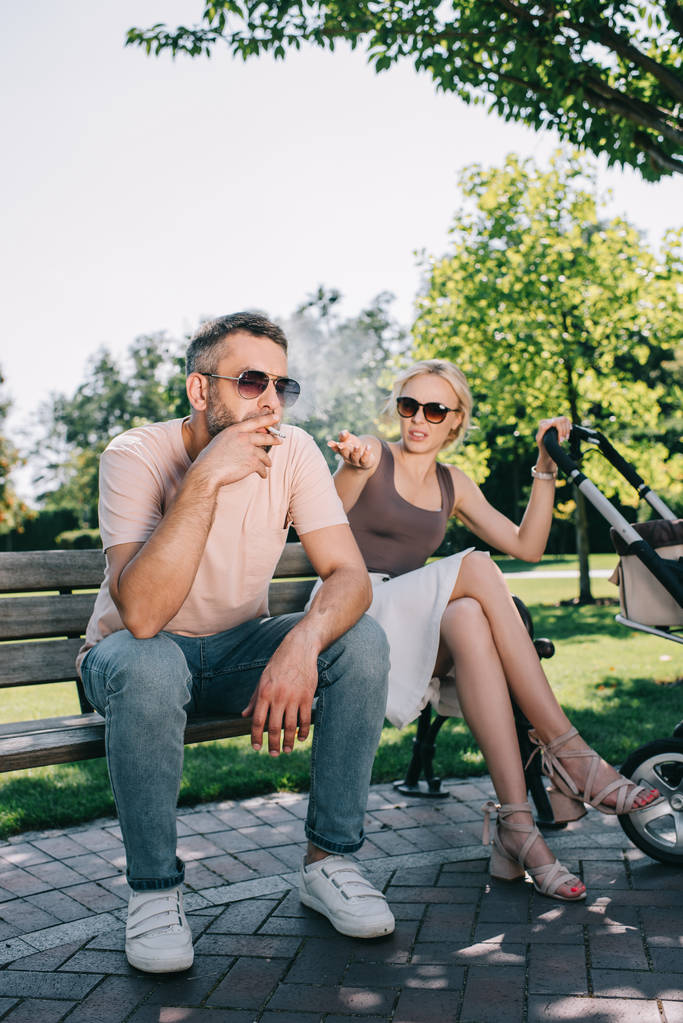 σύζυγος καπνίζοντας τσιγάρο κοντά baby μεταφορά στο πάρκο, σύζυγος χειρονομίες και κοιτάζοντας τον - Φωτογραφία, εικόνα