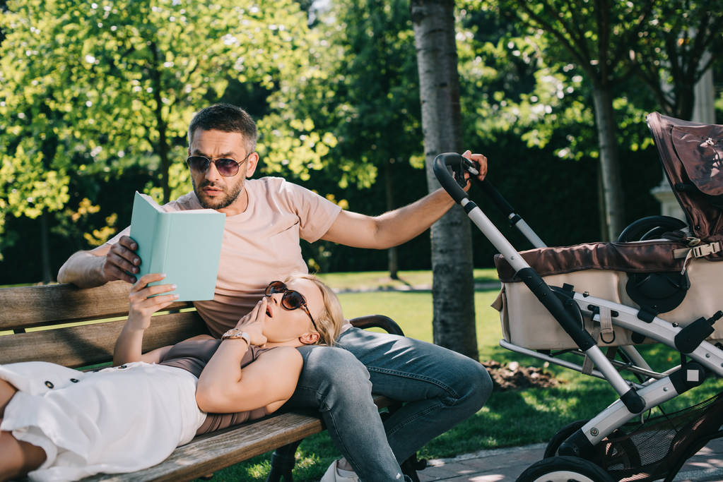 esposa acostada en las piernas del marido cerca de carro de bebé en el parque y libro de celebración, marido leyendo para ella
 - Foto, Imagen