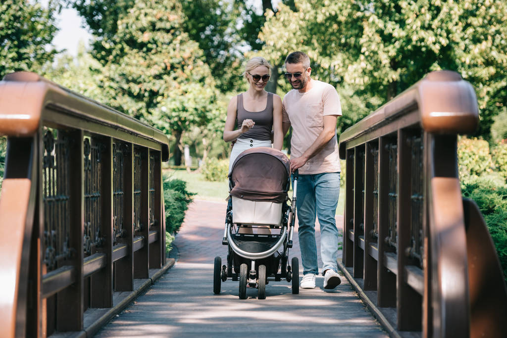 sonrientes padres caminando con carro de bebé en puente en el parque
  - Foto, imagen
