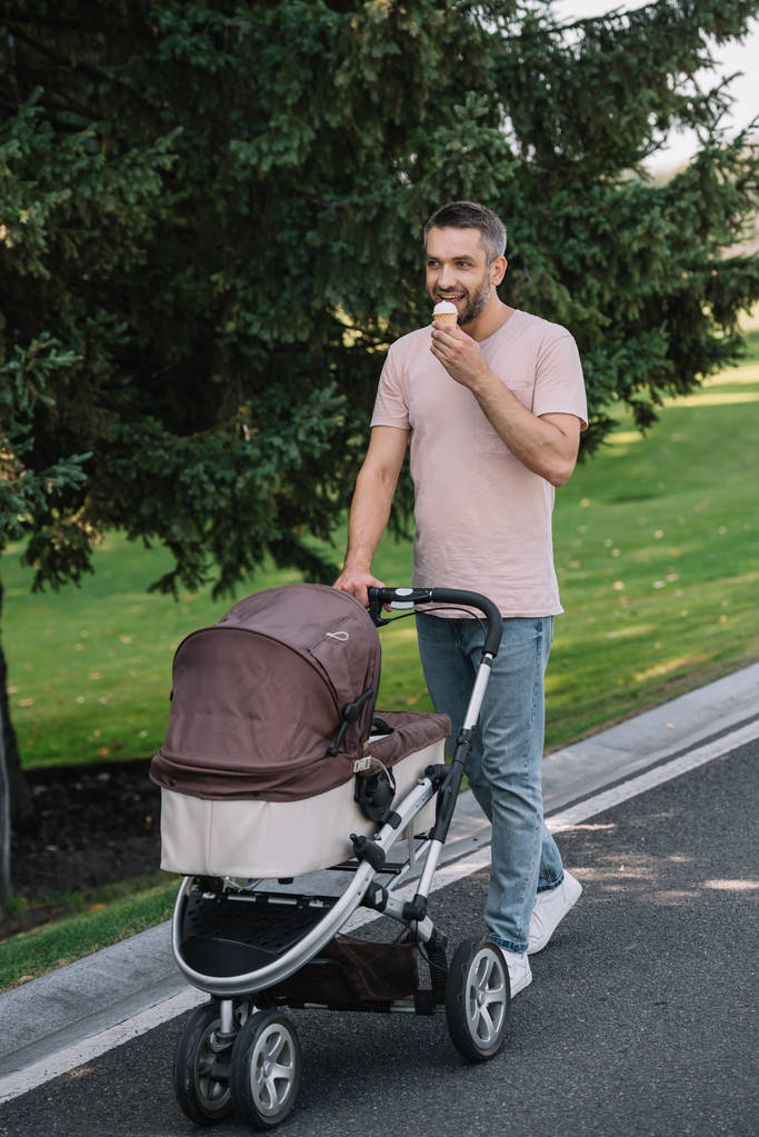 alegre padre caminando con carro de bebé y comiendo helado en el parque
  - Foto, Imagen