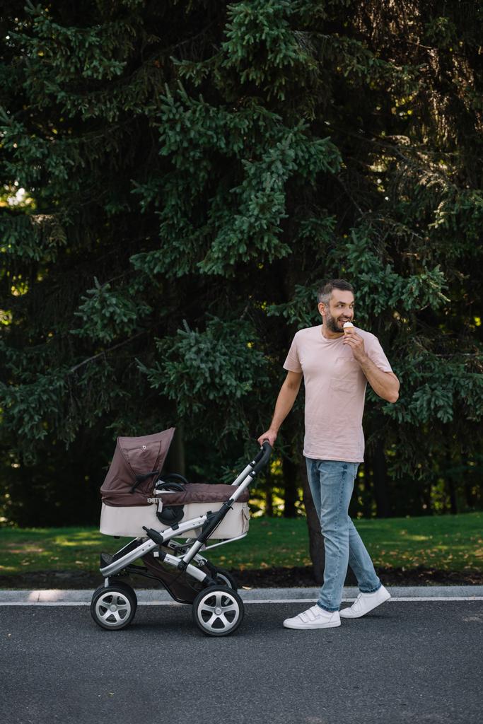 χαμογελά πατέρα περπατώντας με το μωρό μεταφορά και παγωτό στο πάρκο και κοιτάζοντας πίσω - Φωτογραφία, εικόνα