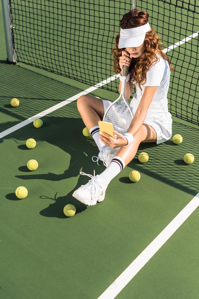 stilvoller Tennisspieler in weißer Sportbekleidung mit Ausrüstung, der am Netz auf dem Tennisplatz ein Selfie mit dem Smartphone macht - Foto, Bild