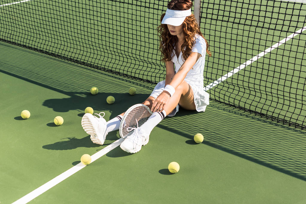 beautiful tennis player in white tennis uniform with racket sitting near tennis net on court with tennis balls around - Photo, Image