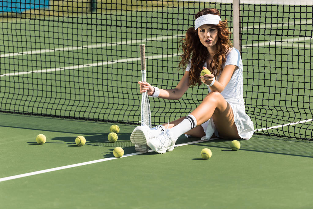 όμορφα γυναικεία αντισφαίριση παίκτης με ρακέτα κάθεται κοντά στο δίχτυ αντισφαίρισης στο Δικαστήριο με μπάλες του τένις στη γύρω περιοχή - Φωτογραφία, εικόνα