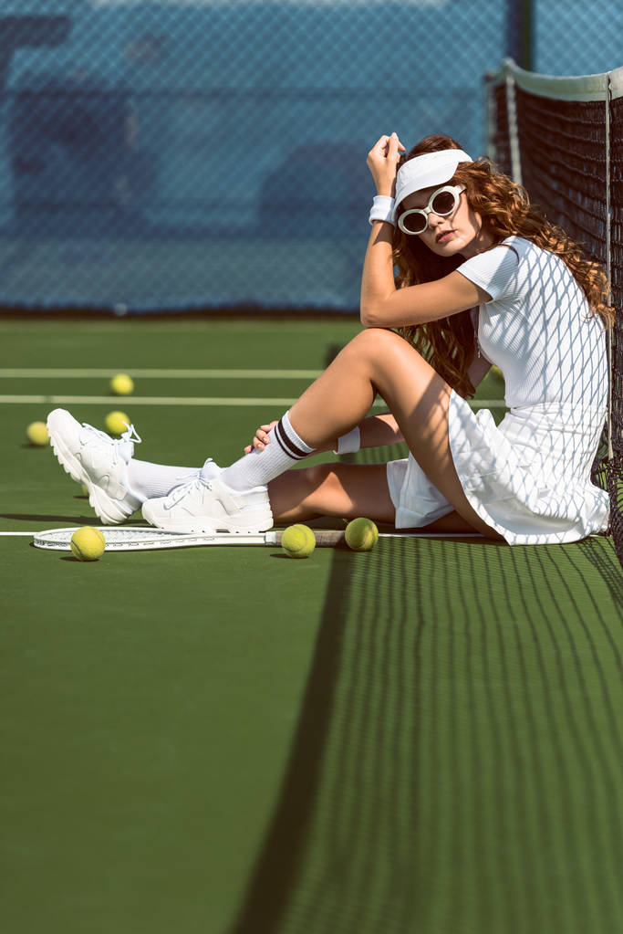 Módní tenistka v sluneční brýle odpočívá nedaleko net na tenisový kurt s zařízení v blízkosti   - Fotografie, Obrázek