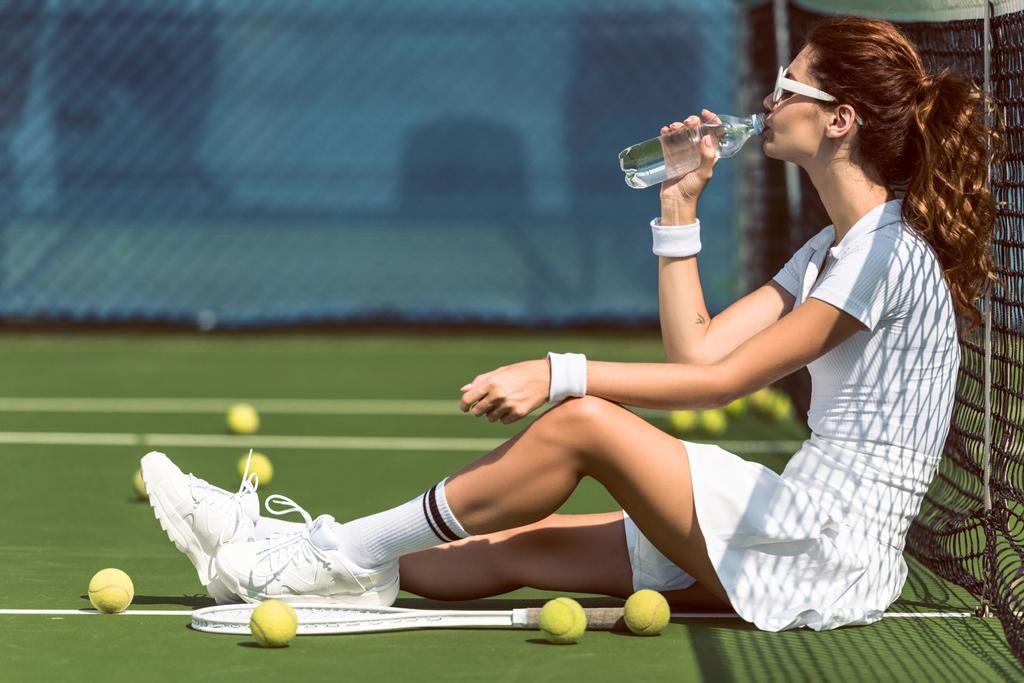 beautiful tennis player in white tennis uniform and sunglasses drinking water while resting on court with racket and balls - Photo, Image