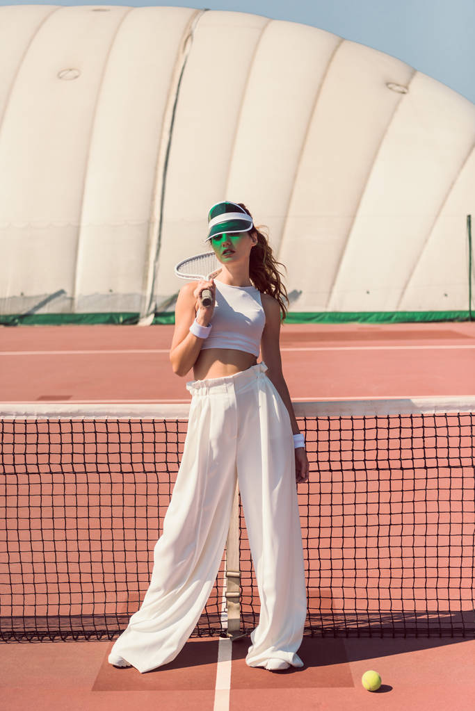 κομψή γυναίκα σε λευκά ρούχα και καπέλο με ρακέτα του τένις που ποζάρουν στο δίχτυ αντισφαίρισης στο γήπεδο - Φωτογραφία, εικόνα