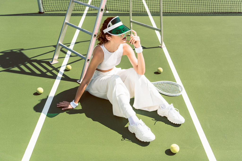 jovem mulher na moda roupas brancas e boné sentado no campo de ténis com raquete e bolas
 - Foto, Imagem