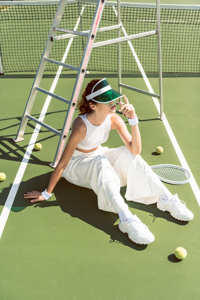 mladá žena v módní bílé oblečení a kryt sedí na tenisový kurt s pálky a míčky - Fotografie, Obrázek