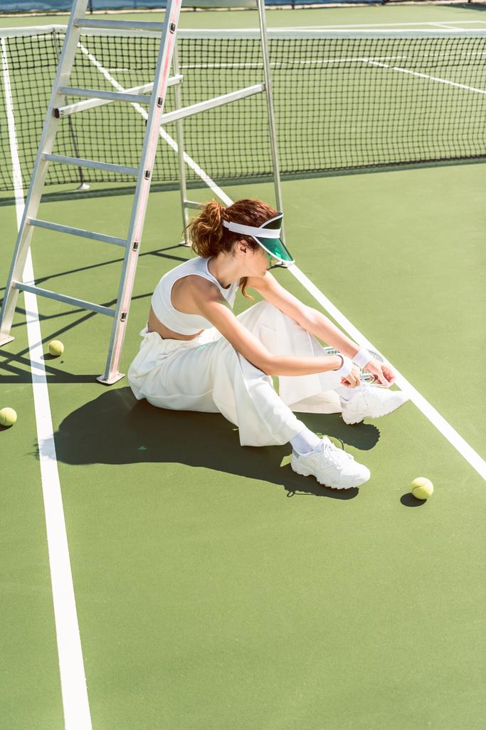 side view of young woman in stylish white clothing and cap tying shoelaces on tennis court with racket and balls - Photo, Image