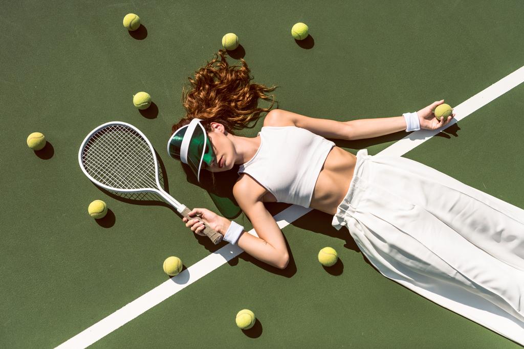 overhead view of stylish woman in white clothing and cap lying with racket lying on tennis court with racket - Photo, Image