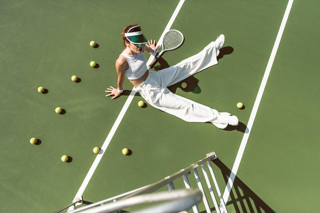 vysoký úhel pohled na krásná žena v elegantní bílé oblečení sedí na tenisový kurt s míčky a povyk kolem - Fotografie, Obrázek
