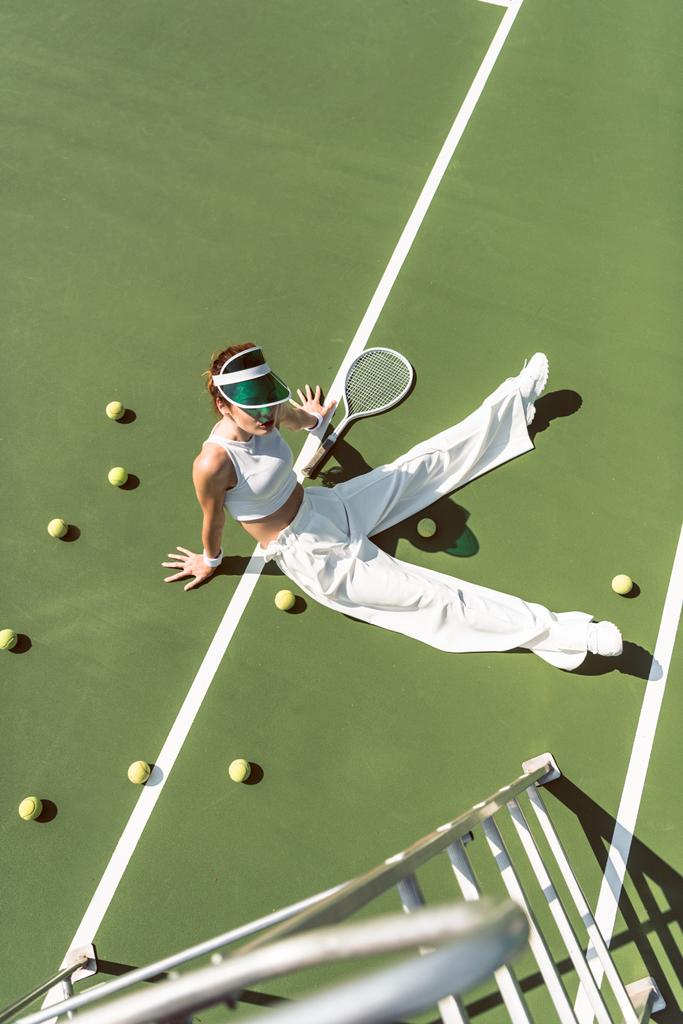 vista de ángulo alto de la hermosa mujer en ropa blanca con estilo sentado en la cancha de tenis con pelotas y raqueta alrededor
 - Foto, imagen