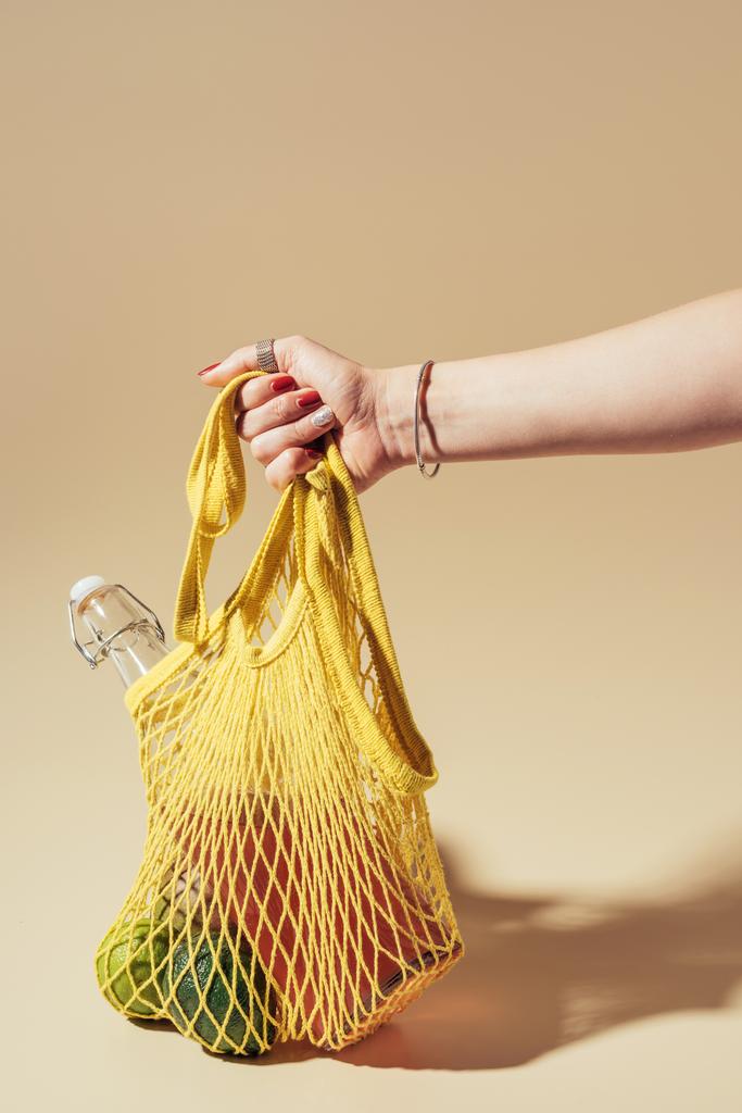 abgeschnittene Aufnahme einer Person mit gelbem Schnurbeutel mit frischem Obst und Glasflasche auf braun  - Foto, Bild