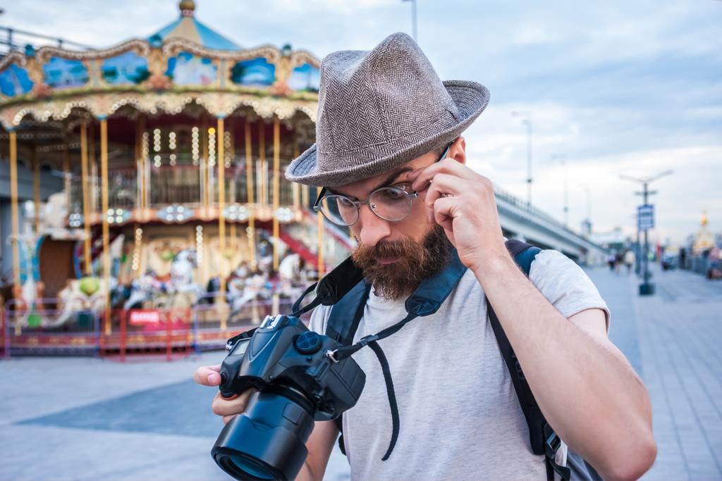 jovem barbudo de chapéu e óculos usando câmera digital no parque de diversões
 - Foto, Imagem