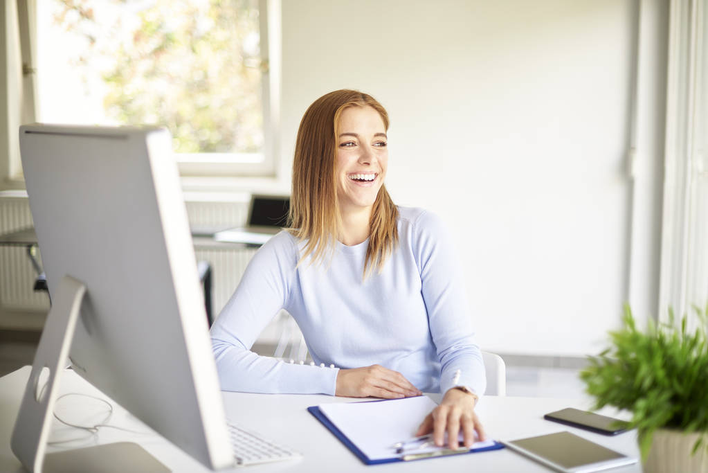 Sonriente joven empresaria sentada frente a la computadora y posando en una oficina luminosa
 - Foto, imagen