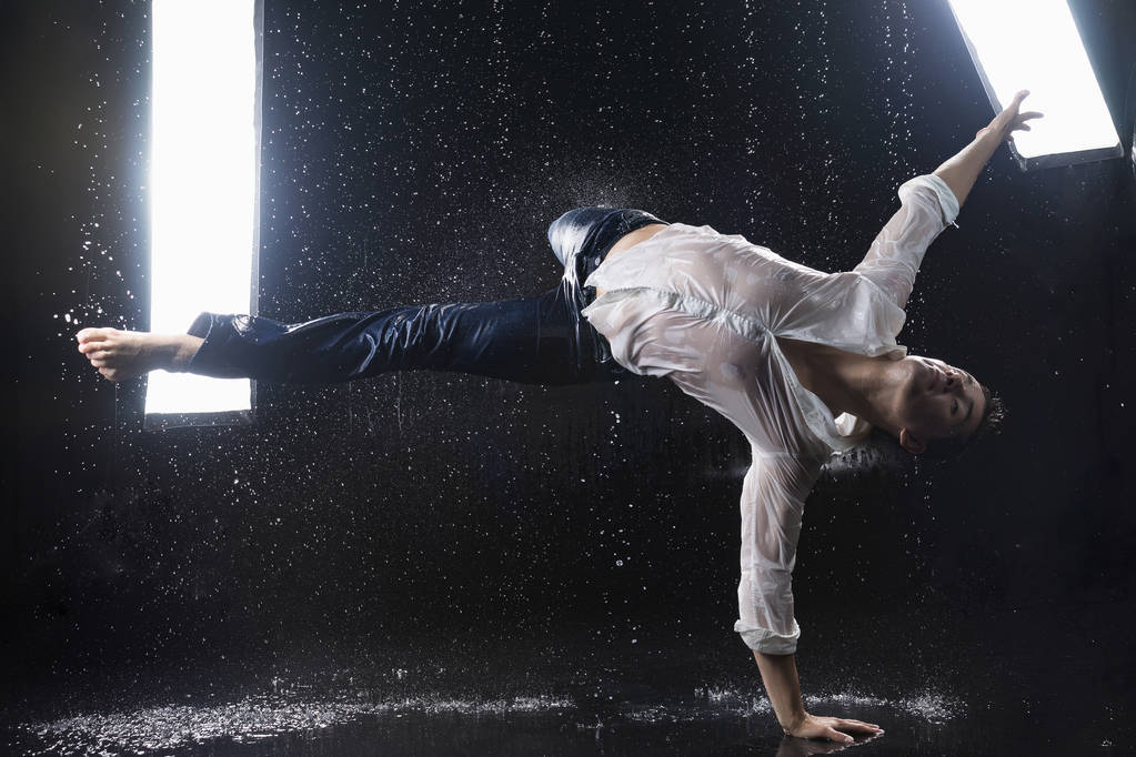 Een barefoot jongeman dragen van natte blauwe spijkerbroek en een wit overhemd cancelbots danst moderne dansen op het water onder regenachtige waterdrops onder de watersplashes. Geïsoleerd op een zwarte achtergrond. Kopiëren van ruimte. - Foto, afbeelding