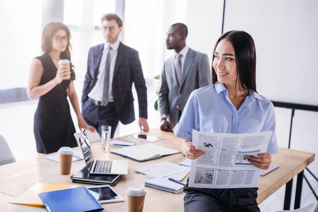 enfoque selectivo de mujer de negocios asiática con periódico y colegas detrás en la oficina
 - Foto, imagen