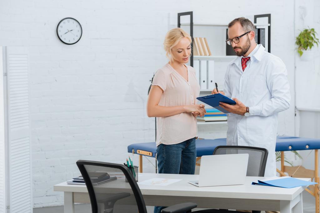 portrait of chiropractic in white coat with notepad and female patient having conversation in clinic - Photo, Image