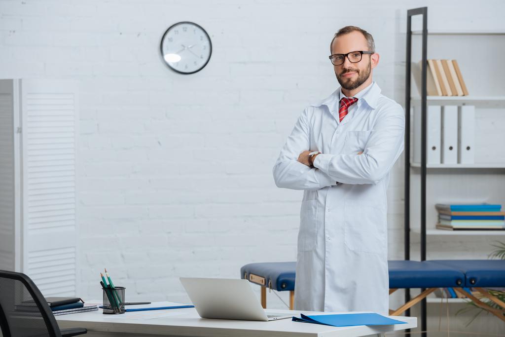 male chiropractor in white coat and eyeglasses with arms crossed standing at workplace with laptop in hospital - Photo, Image
