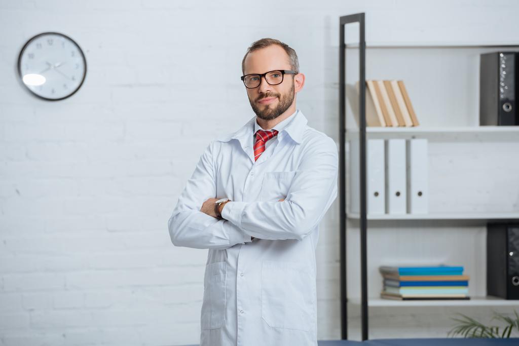 Portret van mannelijke chiropractor in witte jas en bril met armen gekruist in ziekenhuis - Foto, afbeelding