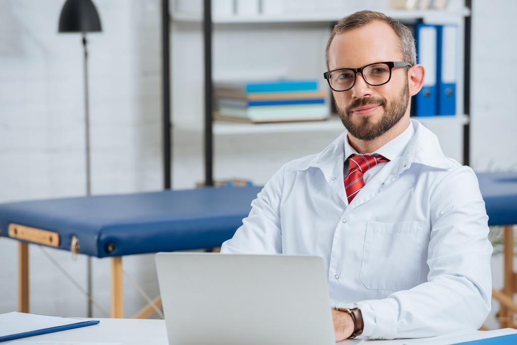 portrait de chiropraticien masculin souriant en manteau blanc sur le lieu de travail avec ordinateur portable à l'hôpital
 - Photo, image