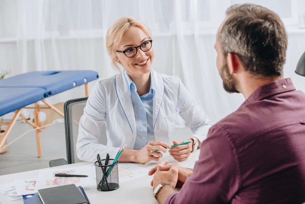 fisioterapista sorridente in camice bianco che guarda il paziente durante l'appuntamento in clinica
 - Foto, immagini