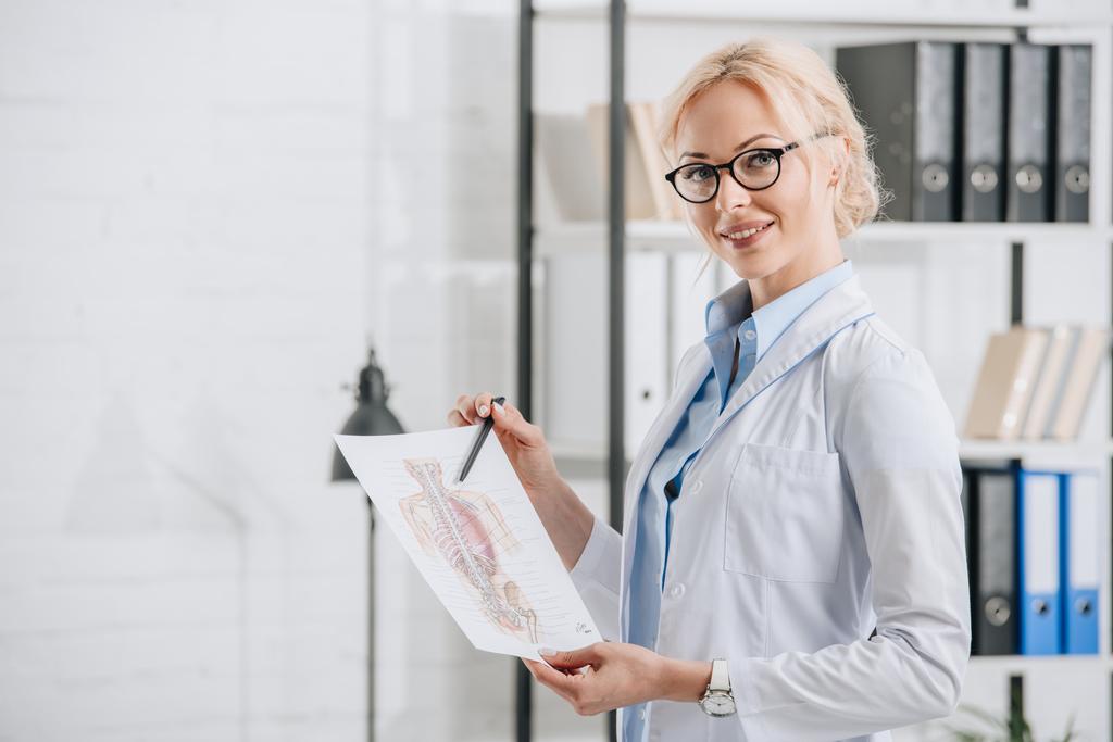 boční pohled na usmívající se chiropraktik v brýle a bílý plášť, ukazuje na lidské tělo schéma v klinice - Fotografie, Obrázek