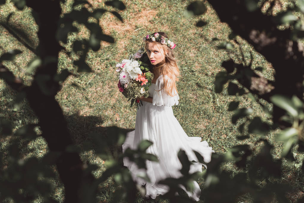 Hochwinkelaufnahme der schönen jungen Braut mit Blumenstrauß und Blick in die Kamera im Freien - Foto, Bild