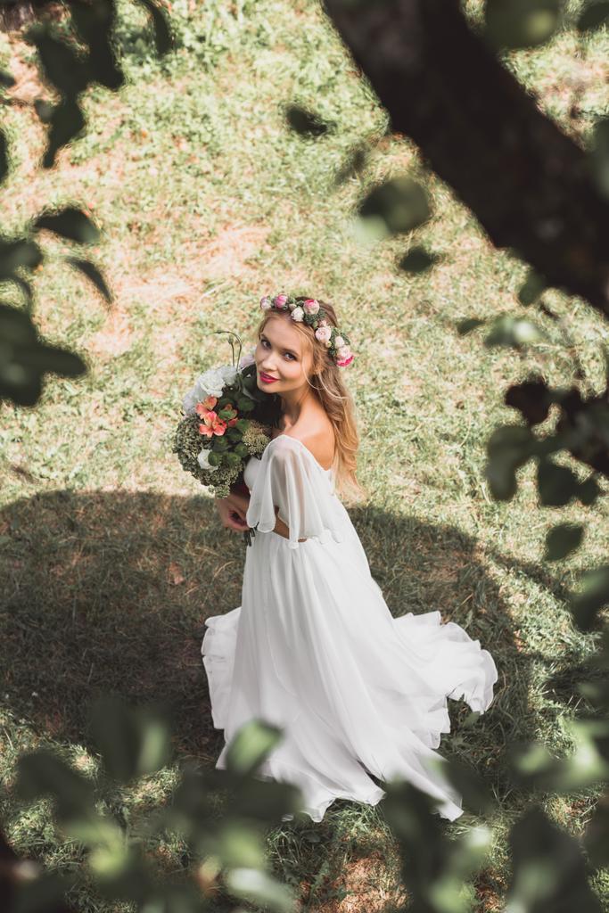 hoge hoekmening van mooie tedere jonge bruid boeket van bloemen en glimlachend in de camera buiten te houden - Foto, afbeelding