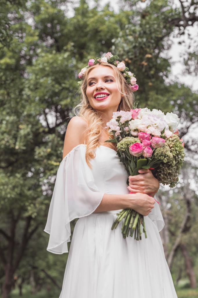 Low-Winkel- Ansicht der lächelnden jungen Braut im Blumenkranz hält Brautstrauß und schaut im Freien weg  - Foto, Bild