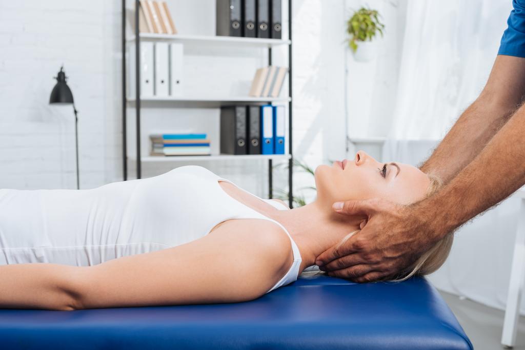 gedeeltelijke weergave van fysiotherapeut masseren van de nek van de patiënt die liggend op een massagetafel in kliniek - Foto, afbeelding
