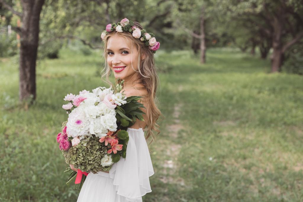 tender young bride in floral wreath and wedding dress holding bouquet of flowers and smiling at camera outdoors   - Photo, Image