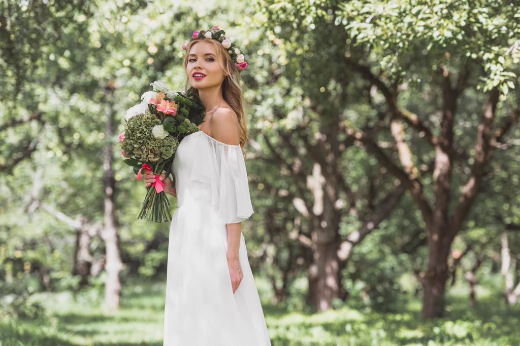 mooie lachende jonge bruid in bloemen krans boeket bloemen houden en kijken naar camera - Foto, afbeelding