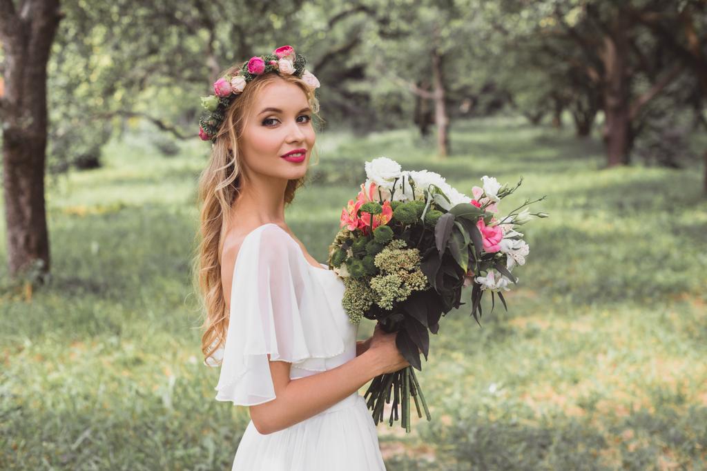 sposa felice in abito da sposa e corona floreale tenendo bouquet di fiori e sorridendo alla fotocamera all'aperto
  - Foto, immagini