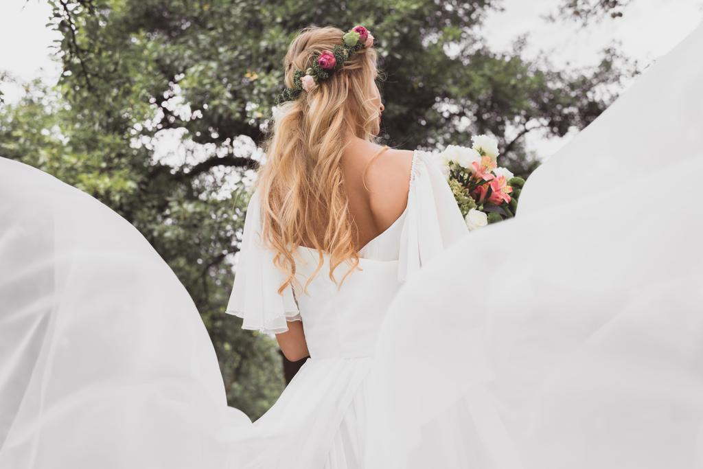 πίσω όψη του νεαρή ξανθιά νύφη στο γάμο φόρεμα εκμετάλλευση μπουκέτο λουλούδια σε εξωτερικούς χώρους - Φωτογραφία, εικόνα