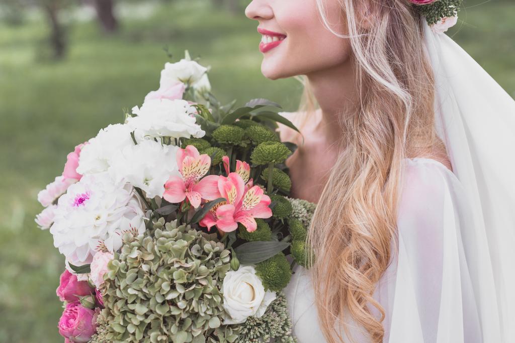 abgeschnittene Aufnahme einer lächelnden blonden Braut mit einem schönen Blumenstrauß im Freien - Foto, Bild