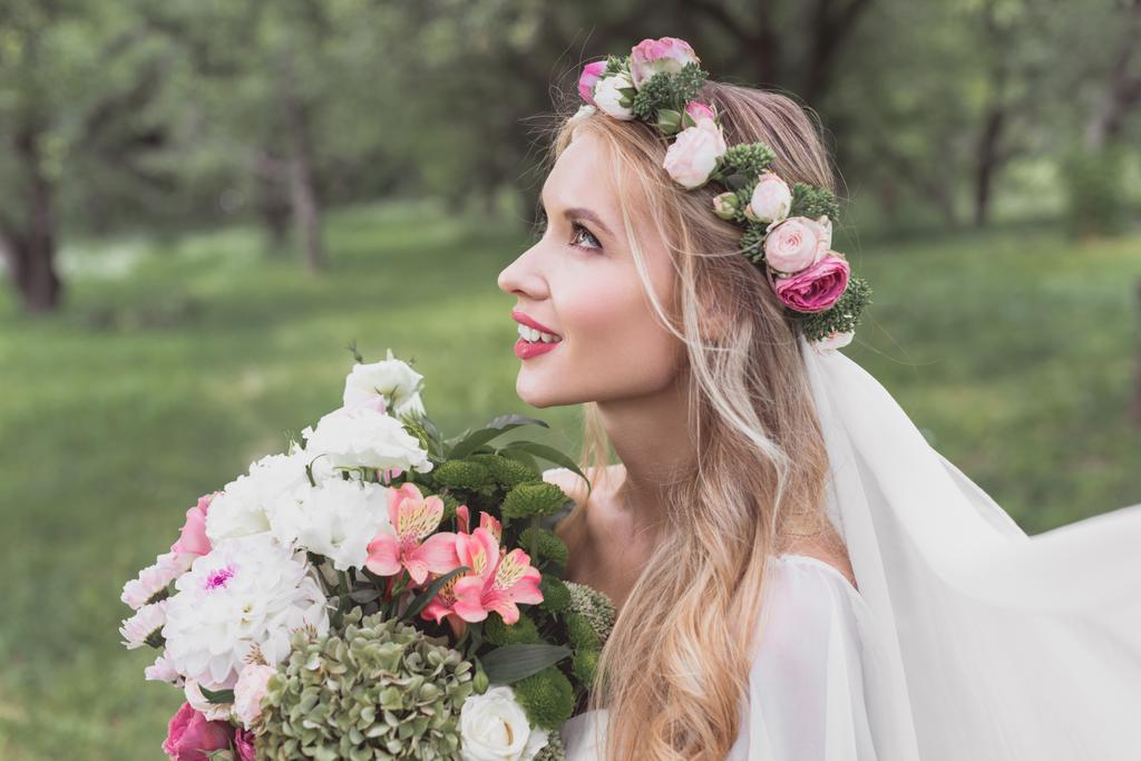side view of beautiful tender bride holding bouquet of flowers and looking up in park - Photo, Image