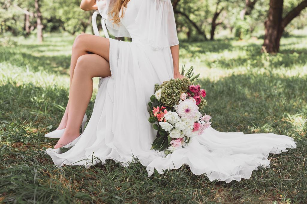 przycięty Zrzut z Panna Młoda w sukni ślubnej, siedząc na krześle i trzymając bukiet kwiatów na zewnątrz - Zdjęcie, obraz