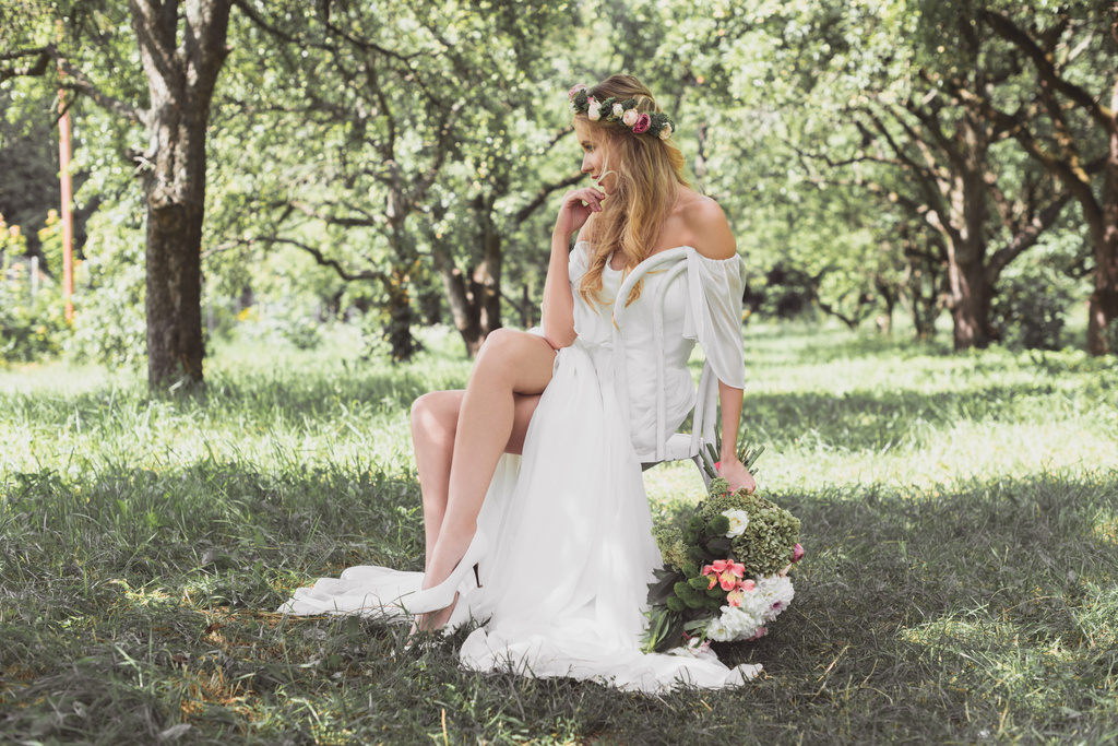 pensive young blonde bride holding wedding bouquet and sitting on chair outdoors  - Photo, Image