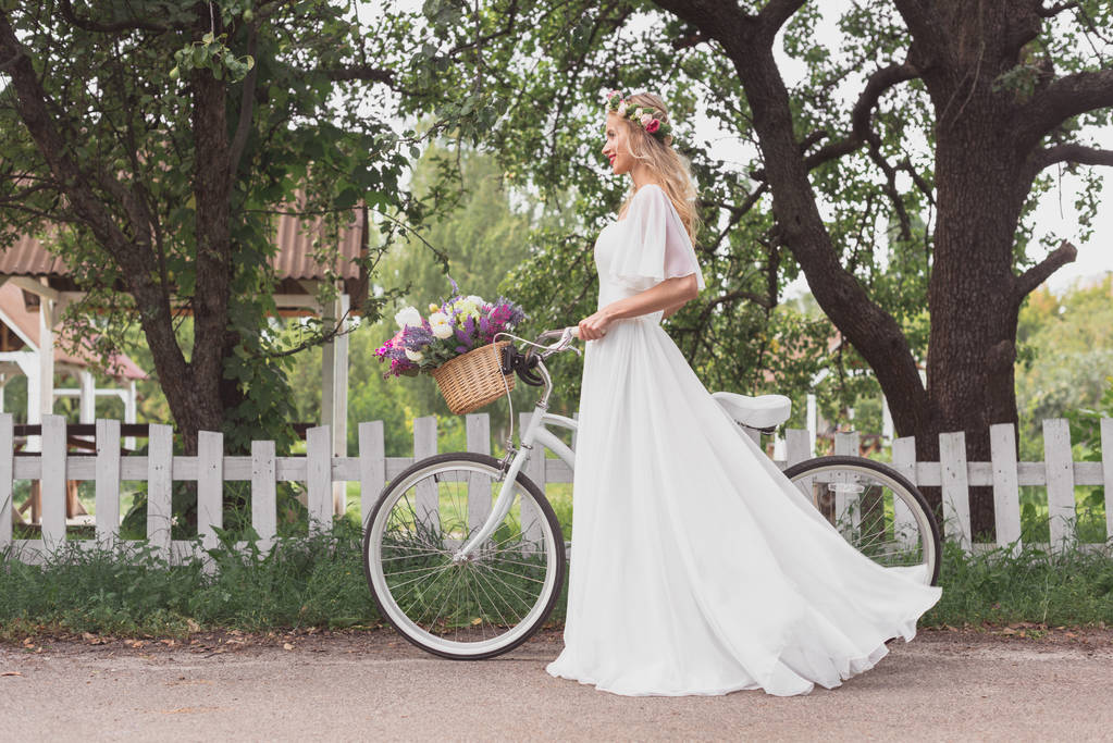 side view of young bride in floral wreath and wedding dress walking with bicycle and looking away - Photo, Image