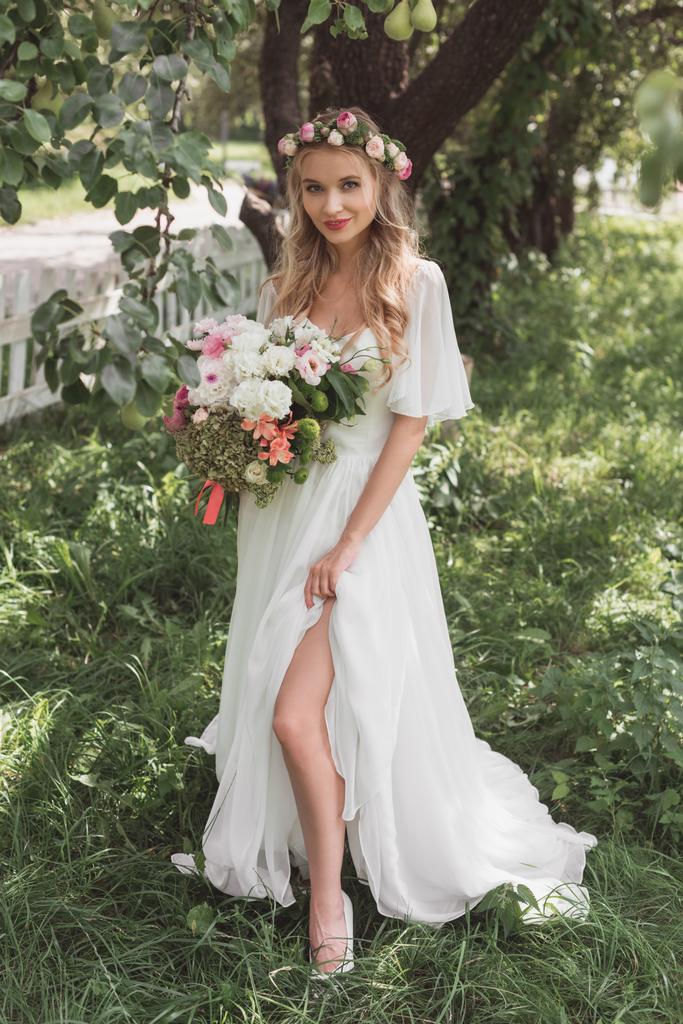 mooie blonde bruid trouwjurk en bloemen krans holding boeket bloemen en glimlachend op camera - Foto, afbeelding