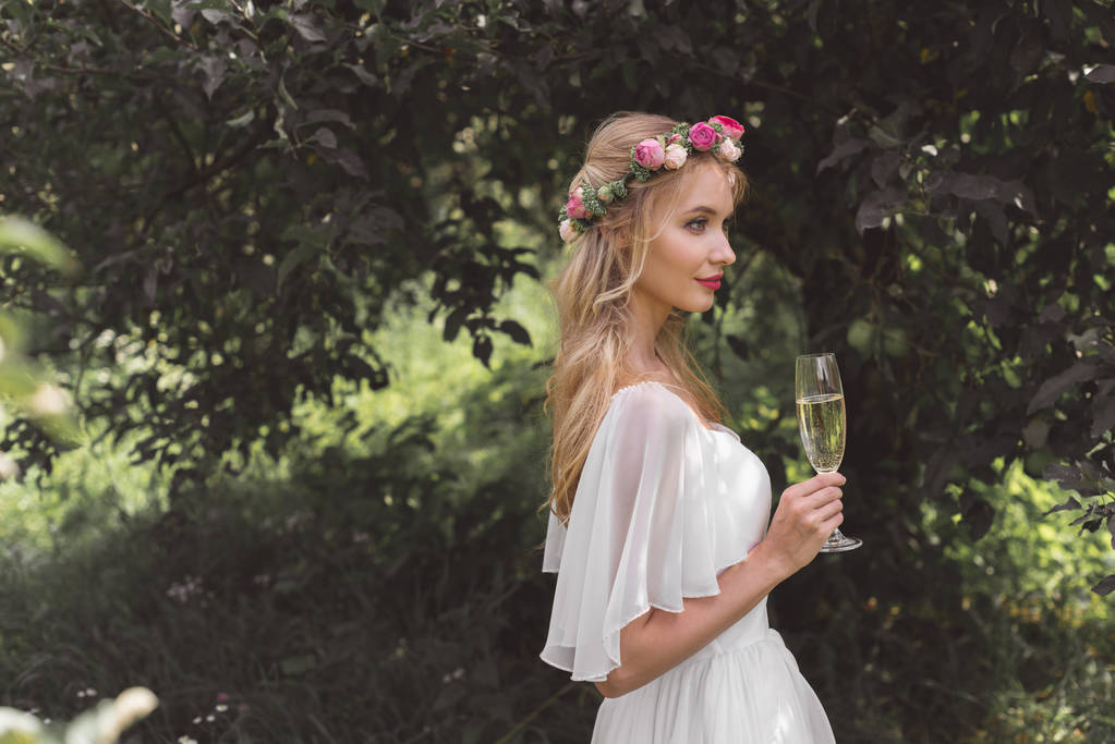 Seitenansicht der schönen jungen Braut im Blumenkranz mit einem Glas Wein im Freien - Foto, Bild