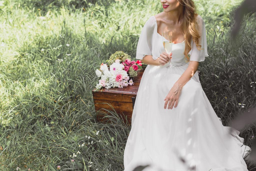 cropped shot of happy young bride holding glass of wine and sitting on vintage chest   - Photo, Image